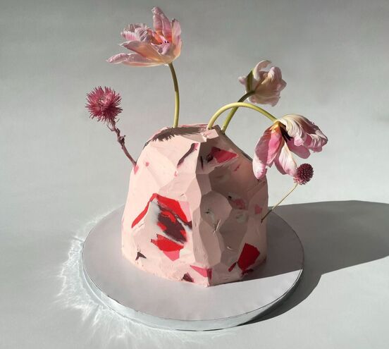 A rock shaped cake iced in pink with red and black areas with fresh pink tulips appearing to sprout from the cake