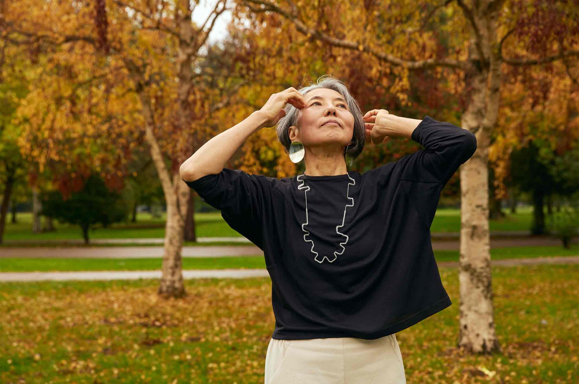 Woman with her hands raised to her hair standing in front of autumnal trees. She is wearing the Asmuss Puzzle Top.