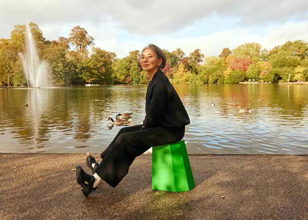 Woman sitting on a bright green Arnold Circus stool in front of a large pond with a fountain and trees in the distance. She is wearing the Asmuss Hepworth Zip Through Sweatshirt and Column Joggers.
